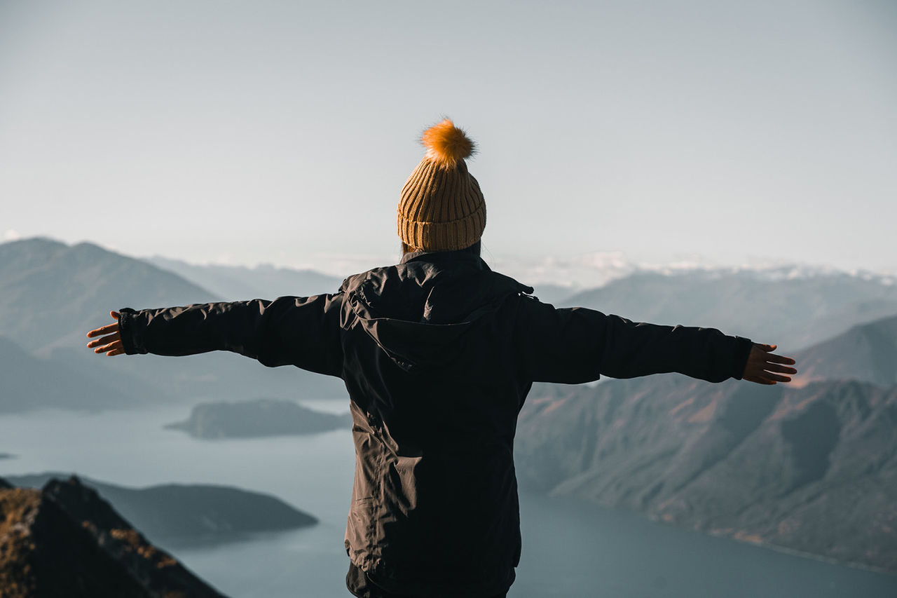 Back view of woman in hat and coat standing on high peak holding hands apart against majestic terrain with lake and mountains